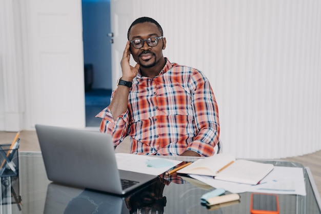 El hombre afroamericano cansado cerró los ojos sosteniendo la cabeza con la mano, tiene un descanso en el lugar de trabajo en la computadora portátil