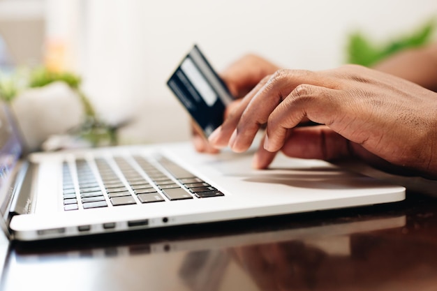 Hombre afroamericano en casa usando una computadora portátil para comprar en línea con tarjeta de crédito en la mano
