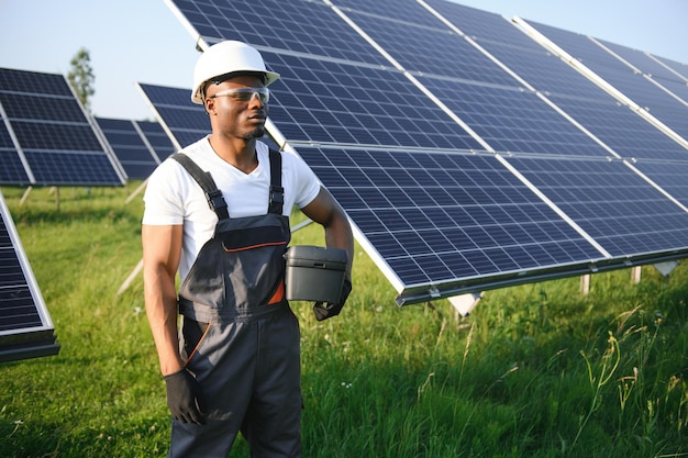 Hombre afroamericano con casco blanco y mono gris parado entre filas de paneles solares