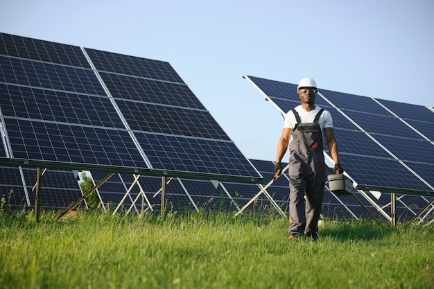 Hombre afroamericano con casco blanco y mono gris de pie entre filas de paneles solares