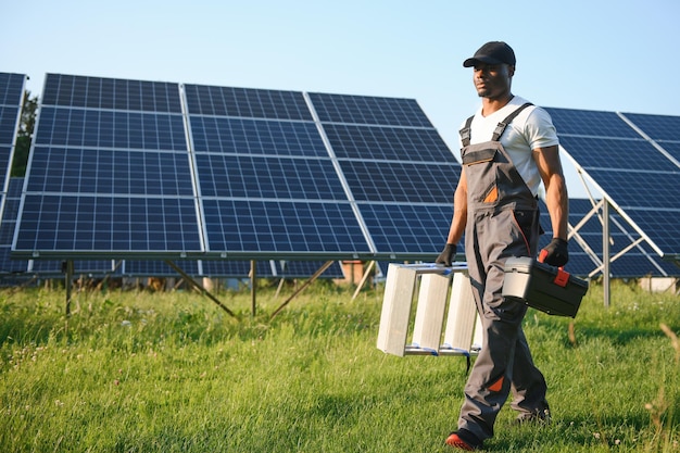 Hombre afroamericano con casco blanco y mono gris de pie entre filas de paneles solares