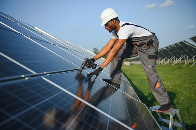 Hombre afroamericano con casco de seguridad y gafas aprieta tuercas en paneles solares con destornillador Técnico competente usando herramientas mientras realiza trabajos de servicio en la estación