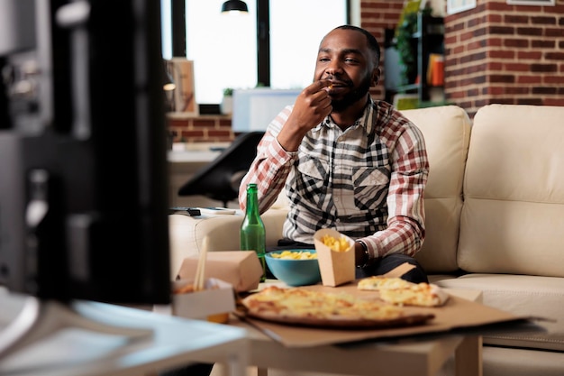 Hombre afroamericano comiendo papas fritas mientras ve películas en la televisión, divirtiéndose con la entrega de comida para llevar y la botella de cerveza. Ver películas en televisión y disfrutar de comida rápida para llevar en casa.