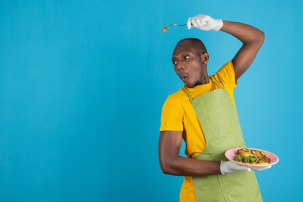 Hombre afroamericano en delantal verde con plato de comida en la pared azul