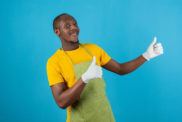 Hombre afroamericano en delantal verde sosteniendo dando pulgar hacia arriba en la pared azul