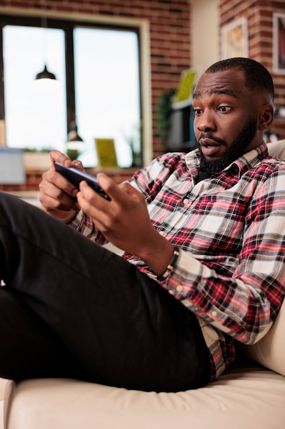 Hombre afroamericano divirtiéndose con videojuegos, jugando competencia en línea en la aplicación de teléfono inteligente. Usando el teléfono móvil y disfrutando de los juegos en Internet, haciendo un trabajo autónomo remoto.