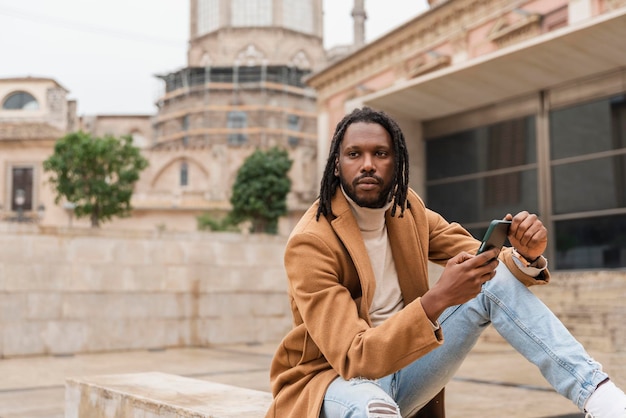 Un hombre afroamericano elegante con rastas sentado en la ciudad al aire libre con un teléfono inteligente en las manos