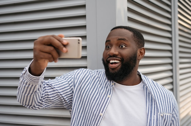 Hombre afroamericano emocionado tomando selfie con teléfono móvil de pie en la calle urbana. Influencer blogger emocional grabando video