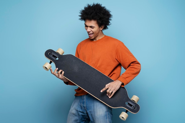 Hombre afroamericano emocional sosteniendo longboard simula tocar guitarra, divirtiéndose, aislado