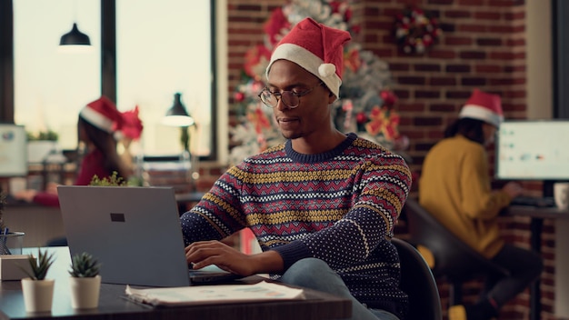 Hombre afroamericano enviando correos electrónicos en una computadora portátil en el escritorio, trabajando en una computadora durante la festividad de la víspera de navidad. Empleado masculino intercambiando ideas antes de celebrar las festivas vacaciones de invierno.