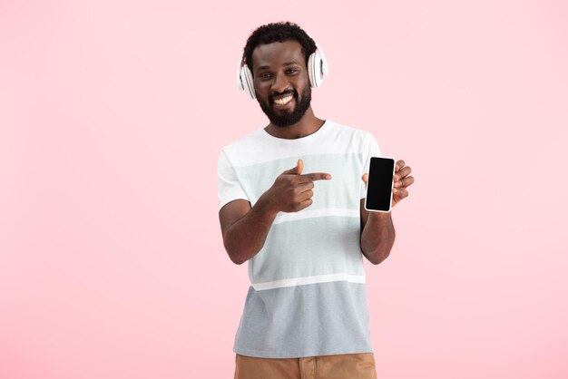 Foto hombre afroamericano escuchando música con auriculares y apuntando al teléfono inteligente con pantalla en blanco