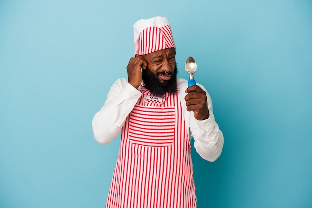 Hombre afroamericano fabricante de helado sosteniendo una bola de helado aislada sobre fondo azul cubriendo las orejas con las manos.