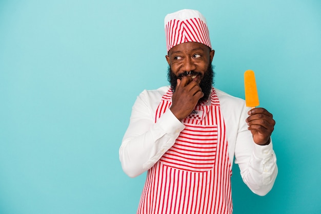 Hombre afroamericano fabricante de helados sosteniendo un helado aislado en la pared azul relajado pensando en algo mirando un espacio de copia.