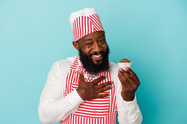 Hombre afroamericano fabricante de helados sosteniendo un helado aislado sobre fondo azul se ríe a carcajadas manteniendo la mano en el pecho.