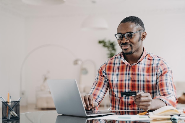 Hombre afroamericano feliz con anteojos está comprando en línea usando una computadora portátil y una tarjeta de crédito