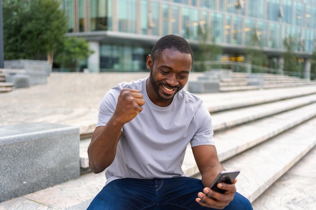 Foto hombre afroamericano feliz ganador eufórico con teléfono inteligente en la calle en la ciudad persona chico mirando