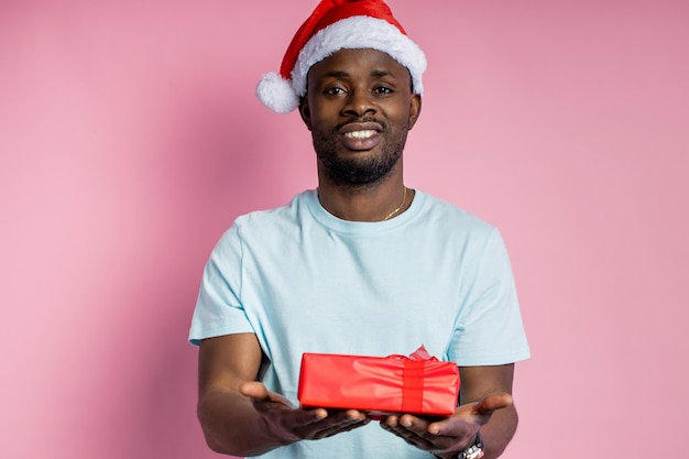 Hombre afroamericano feliz guapo con gorro de Papá Noel, sosteniendo una caja de regalo roja, sonriendo, dando presente a la novia, aislado sobre fondo rosa. Concepto de navidad.