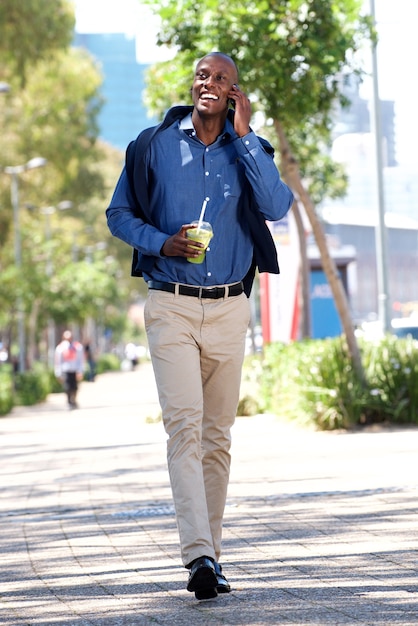 Hombre afroamericano feliz de longitud completa que camina con el teléfono móvil al aire libre