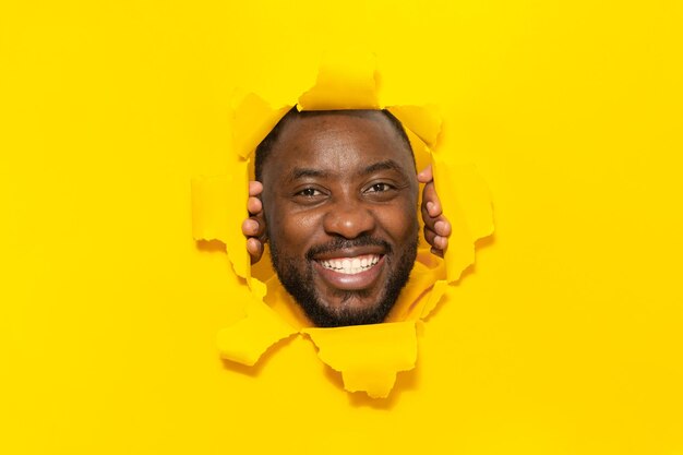 Hombre afroamericano feliz posando en papel amarillo rasgado sonriendo mirando a la cámara a través del agujero en el fondo del estudio