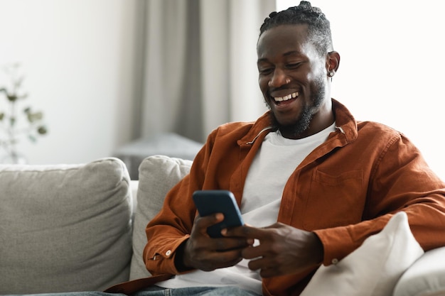 Hombre afroamericano feliz sentado con un teléfono inteligente en las manos y enviando mensajes o navegando en las redes sociales copiando espacio