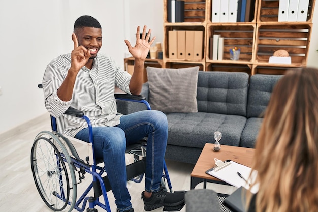 Hombre afroamericano haciendo terapia sentado en silla de ruedas mostrando y señalando con los dedos número seis mientras sonríe confiado y feliz