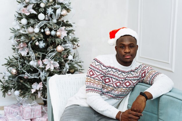 Hombre afroamericano hermoso alegre que mira la cámara y que sonríe interior cerca del árbol adornado que brilla intensamente de la Navidad.