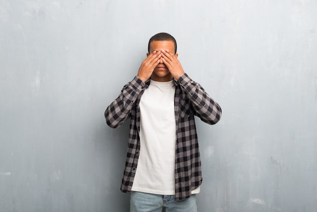 Hombre afroamericano joven con la camisa a cuadros que cubre ojos por las manos