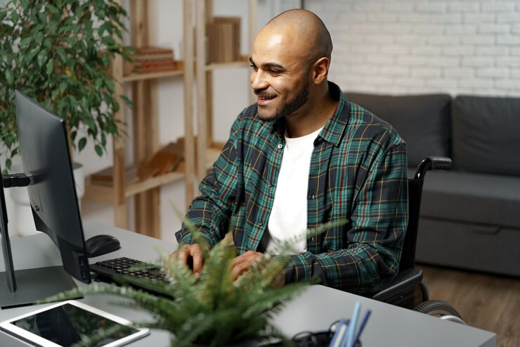 Foto hombre afroamericano joven discapacitado en silla de ruedas usando la computadora mientras está sentado en su mesa de trabajo