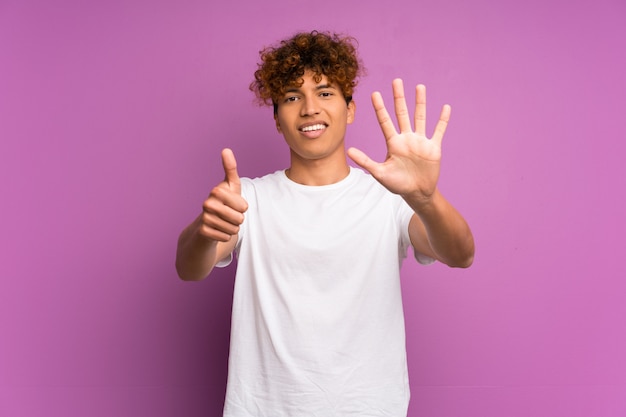 Hombre afroamericano joven sobre la pared púrpura aislada que cuenta seis con los dedos