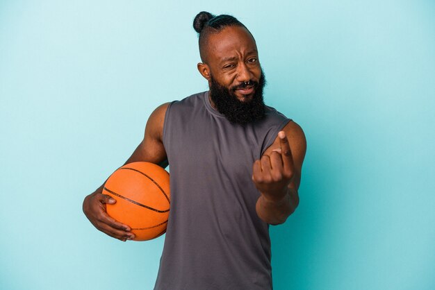 Hombre afroamericano jugando baloncesto aislado sobre fondo azul apuntando con el dedo como si invitara a acercarse.