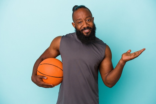 Hombre afroamericano jugando baloncesto aislado sobre fondo azul que muestra un espacio de copia en una palma y sosteniendo otra mano en la cintura.