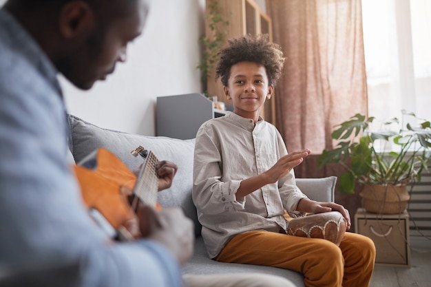 Hombre afroamericano maduro y su hijo adolescente pasar un buen rato juntos en casa tocando música con guitarra y djembé