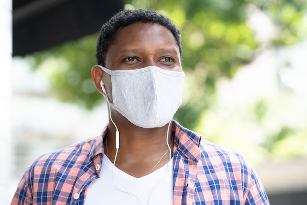 Hombre afroamericano con una mascarilla mientras escucha música con auriculares al aire libre en la calle. Nuevo concepto de estilo de vida normal.