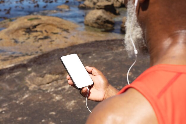 Hombre afroamericano mayor que usa auriculares usando un teléfono inteligente junto al mar. jubilación saludable tecnología comunicación fitness al aire libre estilo de vida.