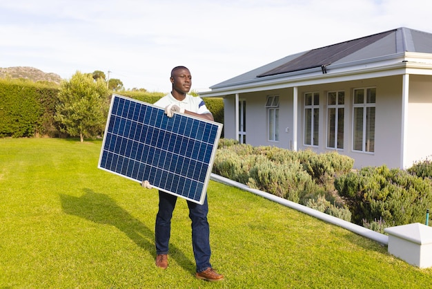 Hombre afroamericano de mediana edad que lleva un panel solar mientras camina sobre tierra verde en el patio contra el cielo