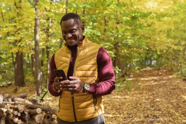 Hombre afroamericano milenario caminando por la calle de la ciudad parque de otoño sosteniendo un teléfono móvil para bloguear co