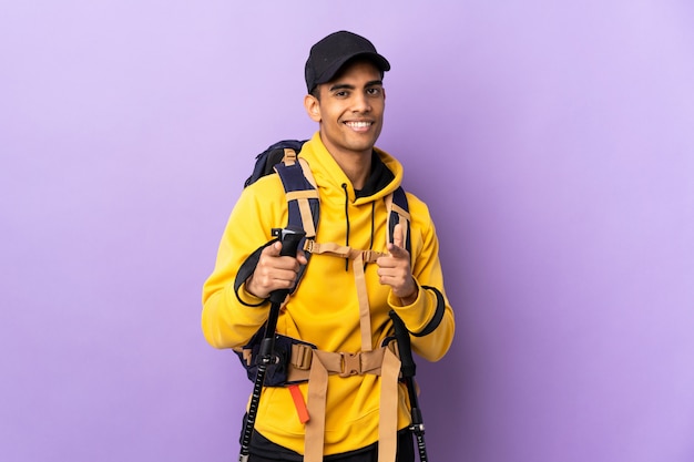 Hombre afroamericano con mochila y bastones de trekking sobre pared aislada apuntando hacia el frente y sonriendo