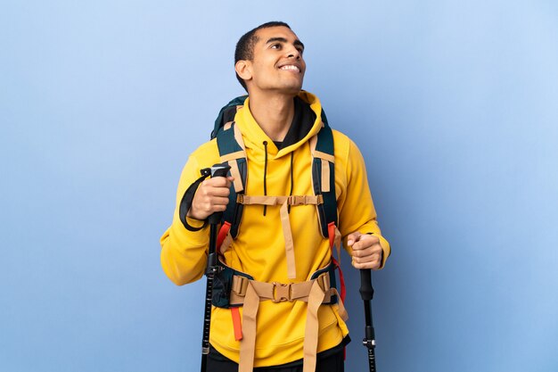 Foto hombre afroamericano con mochila y bastones de trekking sobre pared aislada mirando hacia arriba mientras sonríe