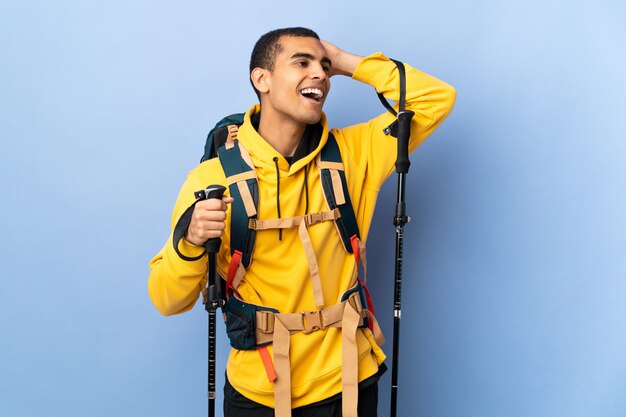 Hombre afroamericano con mochila y bastones de trekking sobre pared aislada sonriendo mucho