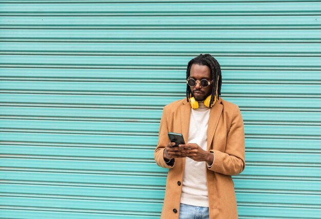 Hombre afroamericano moderno con elegantes gafas de sol enviando un mensaje con teléfono móvil aislado en fondo azul