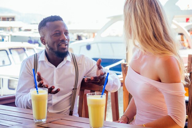 Un hombre afroamericano y una mujer se miran con amor cada uno en un café tropical de verano y beben un cóctel de frutas frescas y naranjas.