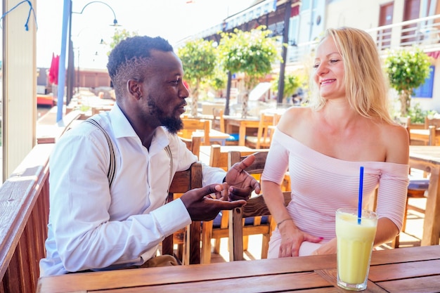 Un hombre afroamericano y una mujer se miran con amor en un café tropical de verano y beben un cóctel de frutas frescas y naranjas