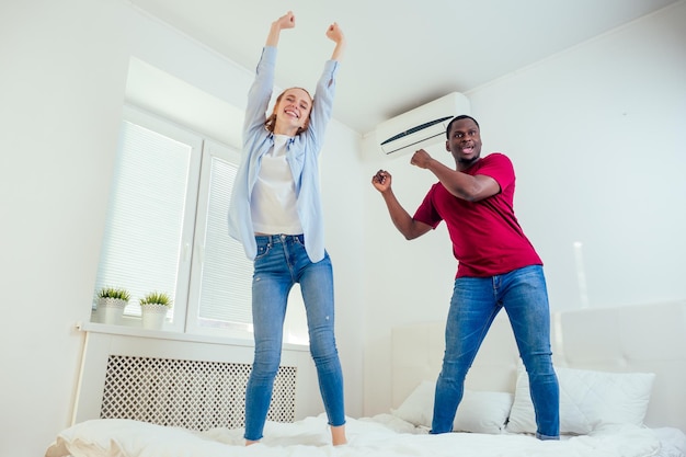 Hombre afroamericano y mujer pelirroja pelirroja riéndose y saltando en la cama en la habitación del resort.