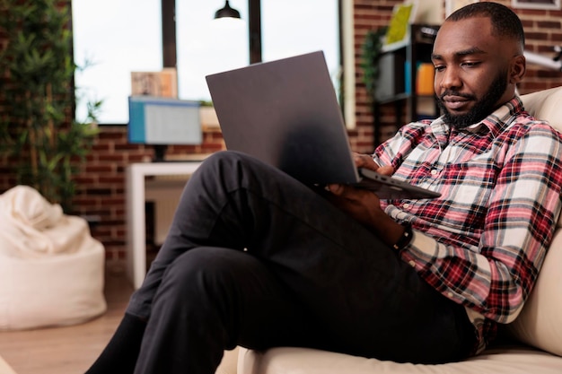 Hombre afroamericano navegando por internet en una laptop, trabajando remotamente desde casa para encontrar información de investigación. Haciendo freelance remoto en la computadora, creando un informe de presentación y sentado en el sofá.