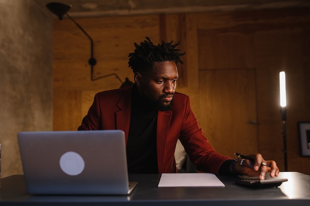 Hombre afroamericano negro guapo que trabaja en la computadora portátil mientras está sentado detrás del escritorio en la acogedora sala de estar independiente trabajando desde casa