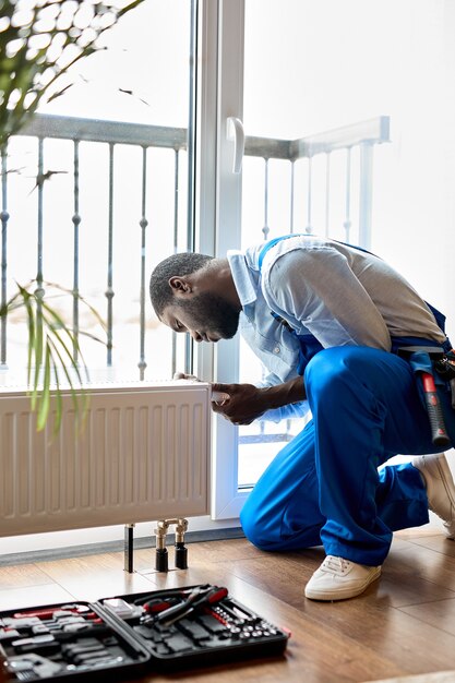 Hombre afroamericano en overoles de ropa de trabajo con herramientas mientras se instala el radiador de calefacción en la habitación