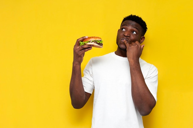 hombre afroamericano pensativo con camiseta blanca sosteniendo una gran hamburguesa y soñando despierto