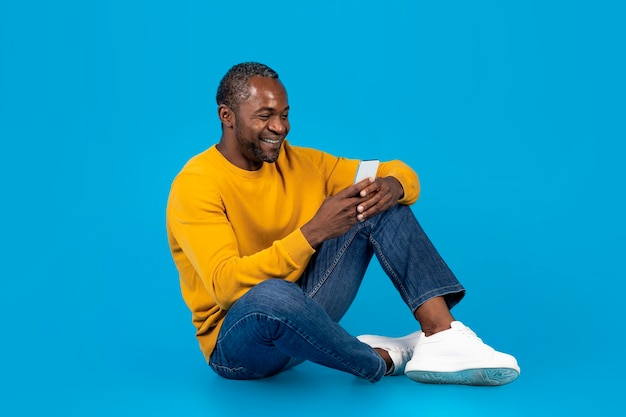 Hombre afroamericano positivo sentado en el suelo usando un teléfono inteligente