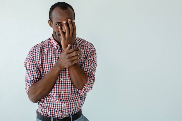 Hombre afroamericano positivo sosteniendo su mano en la cara mientras está de pie contra la pared blanca