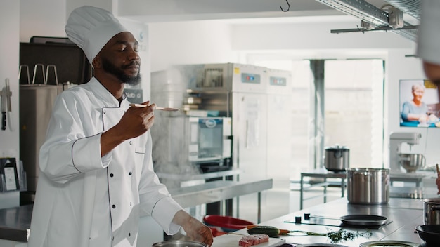 Hombre afroamericano prueba sopa de verduras en la estufa, preparando recetas gastronómicas con ingredientes deliciosos. Auténtico cocinero haciendo salsa y plato culinario fresco, restaurante de comida.